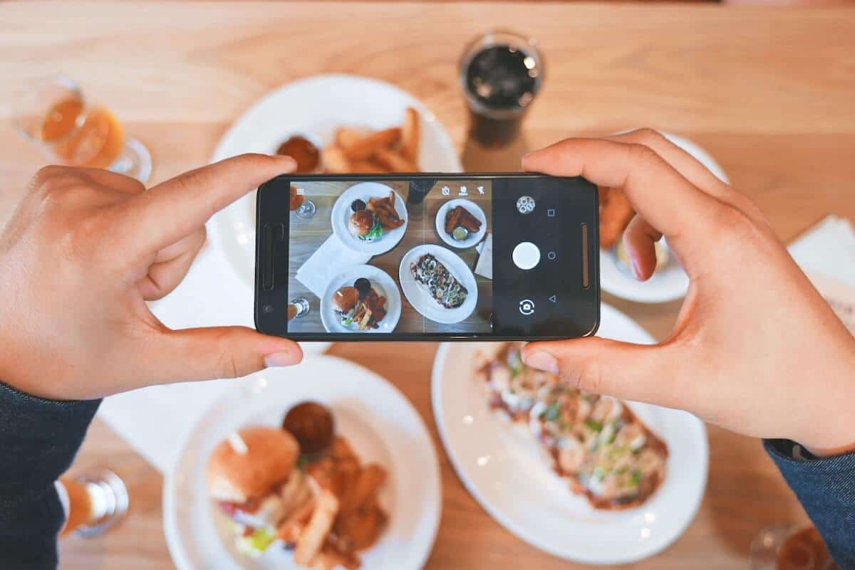 Cover photo for the Best Apollo Bay Restaurants, Bars & Cafes featuring 4 dishes on the table with a woman holding a mobile phone above to take a flatlay photo with the focus on the phone screen