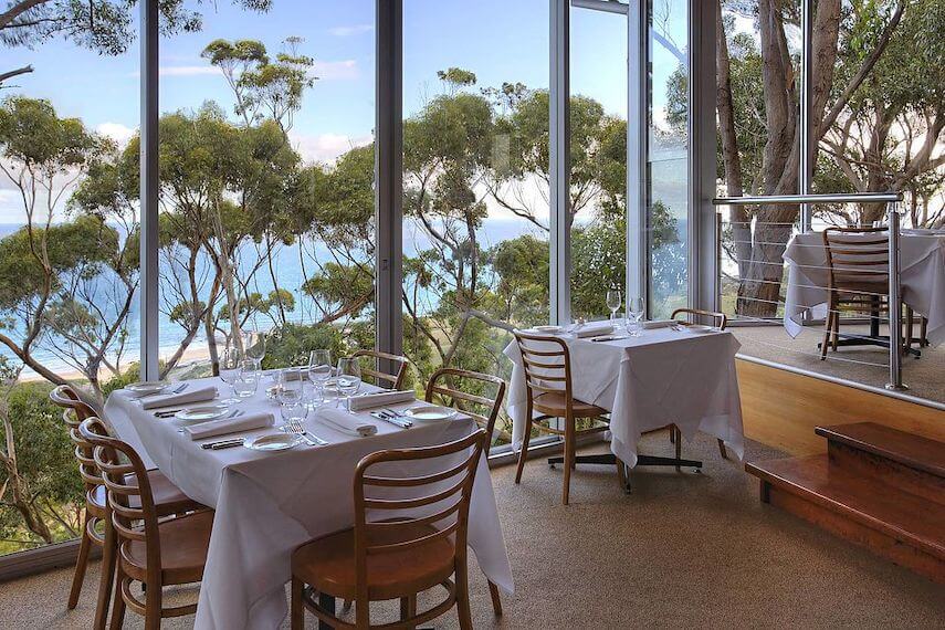 Interior of Chriss' Restaurant looking out over two wooden tables covered in white table cloths next to floor to ceiling windows overlooking the green treetops and blue ocean beyond