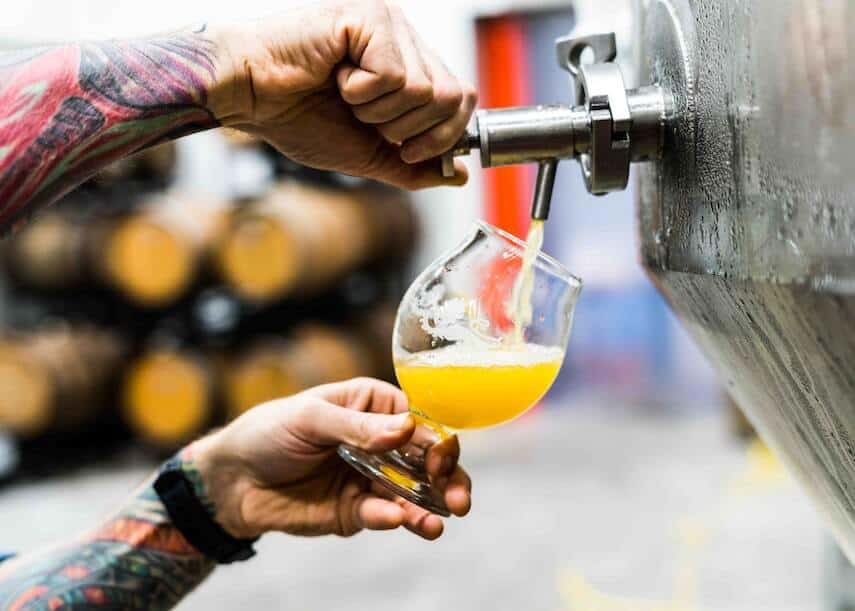 Close up shot of a mans tattooed arms and hands holding a pint pot on an angle while dispensing beer from a stainless steel keg