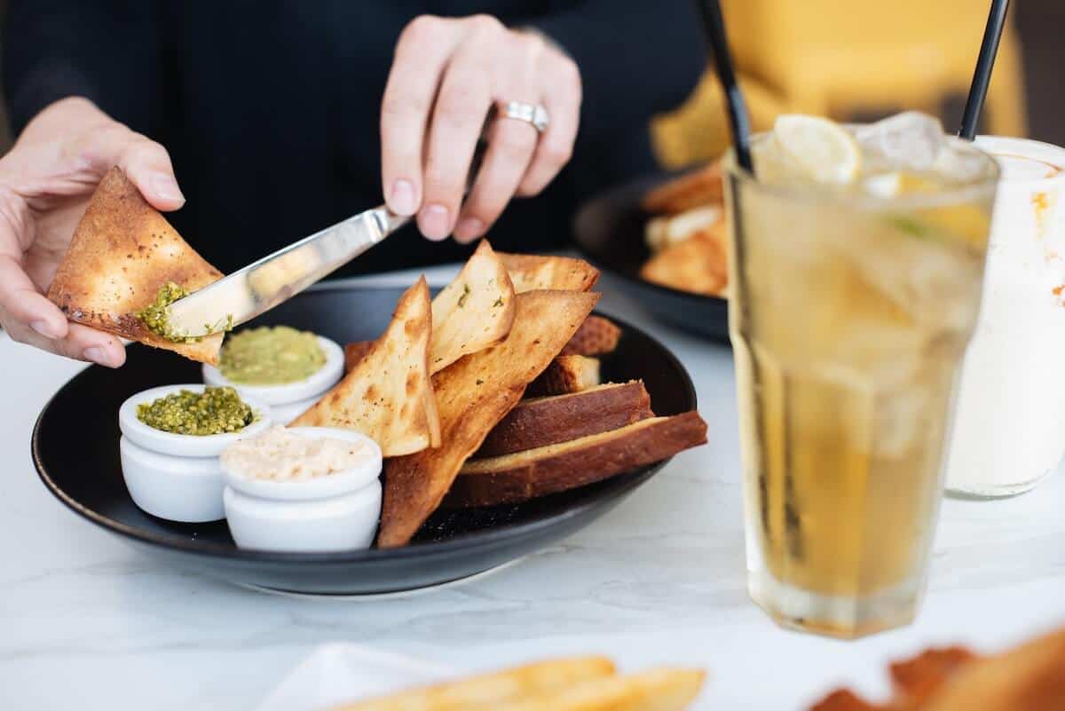 Top Torquay Restaurants cover photo of a man sitting at a table from the chest down putting dip and dukkah onto a slice of toasted bread, and a glass of ice tea with ice