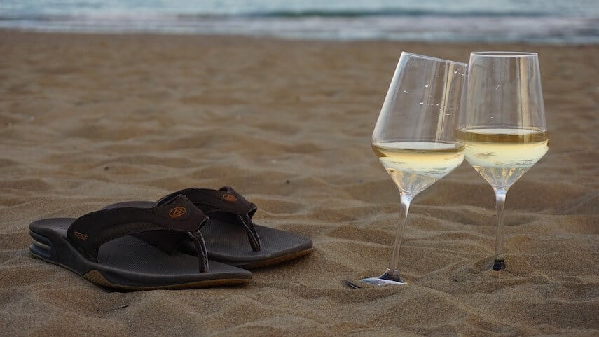 Two glasses of wine propped against each other on the beach next to a brown pair of flip flops with the ocean in the background