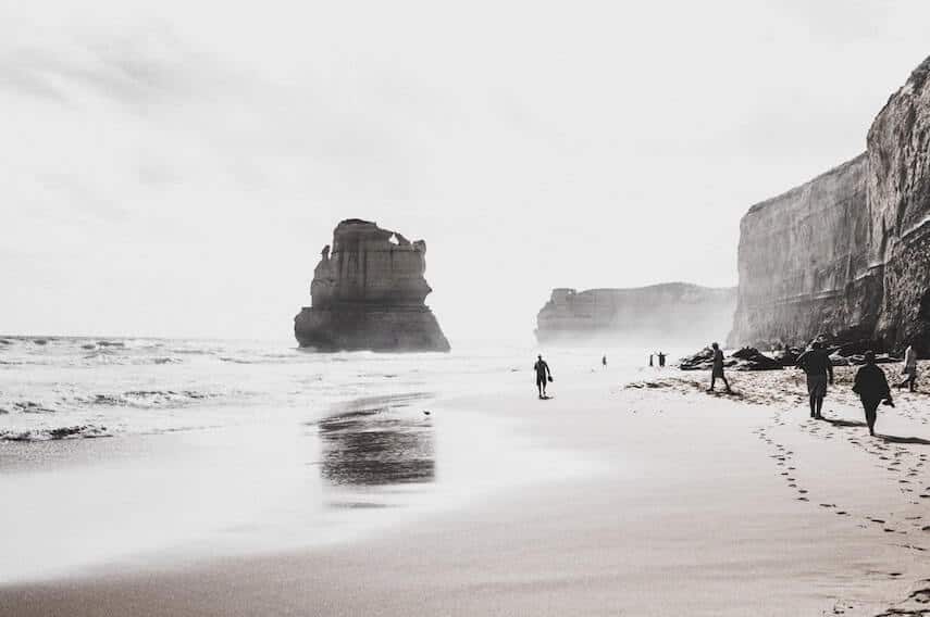 Cover photo for 5 Day Great Ocean Road Itinerary featuring Black and white image of the beach at Gibson Steps with a large limestone rock standing tall in the ocean and people walking on the sand towards it