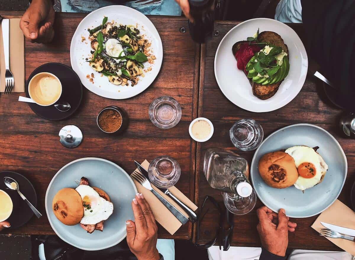 Cover photo for the Best Anglesea Restaurants, Bars & Cafes featuring a top-down shot of 4 plates of food on a table along with knives and forks, water glasses and salt and pepper shaker