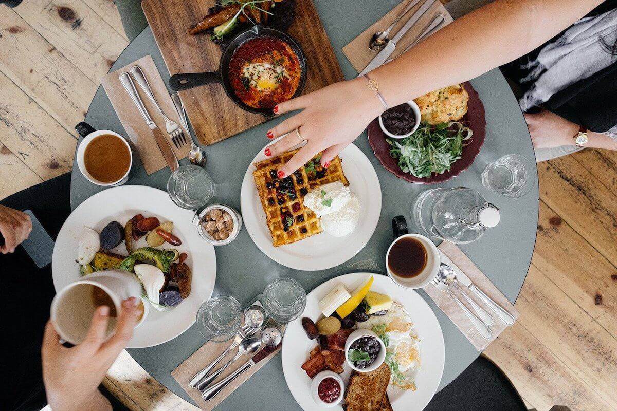 Cover photo for the Best Port Campbell Restaurants, Bars & Cafes featuring a top down shot of a table with 4 dishes on plates and 1 dish on a wooden board with a womans hand with red nail varnish reaching into the middle of the table