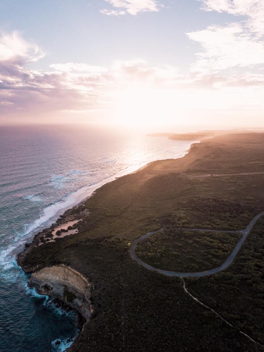 Great Ocean Road Itinerary Cover Shot featuring an aerial shot of a road winding along the cliff edge with the ocean below and the sunset in the background