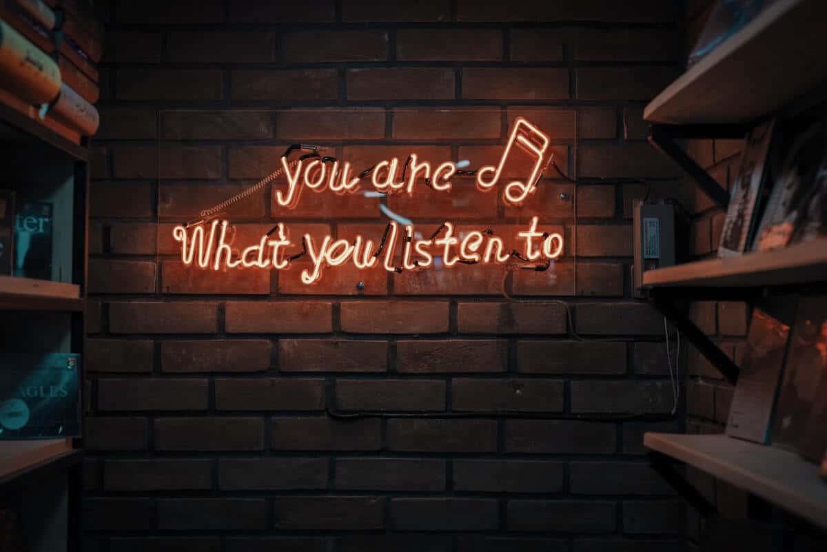 Neon sign on a brick wall: "you are what you listen to" - cover photo for the 100 Best Travel Songs for the Ultimate Travel Playlist