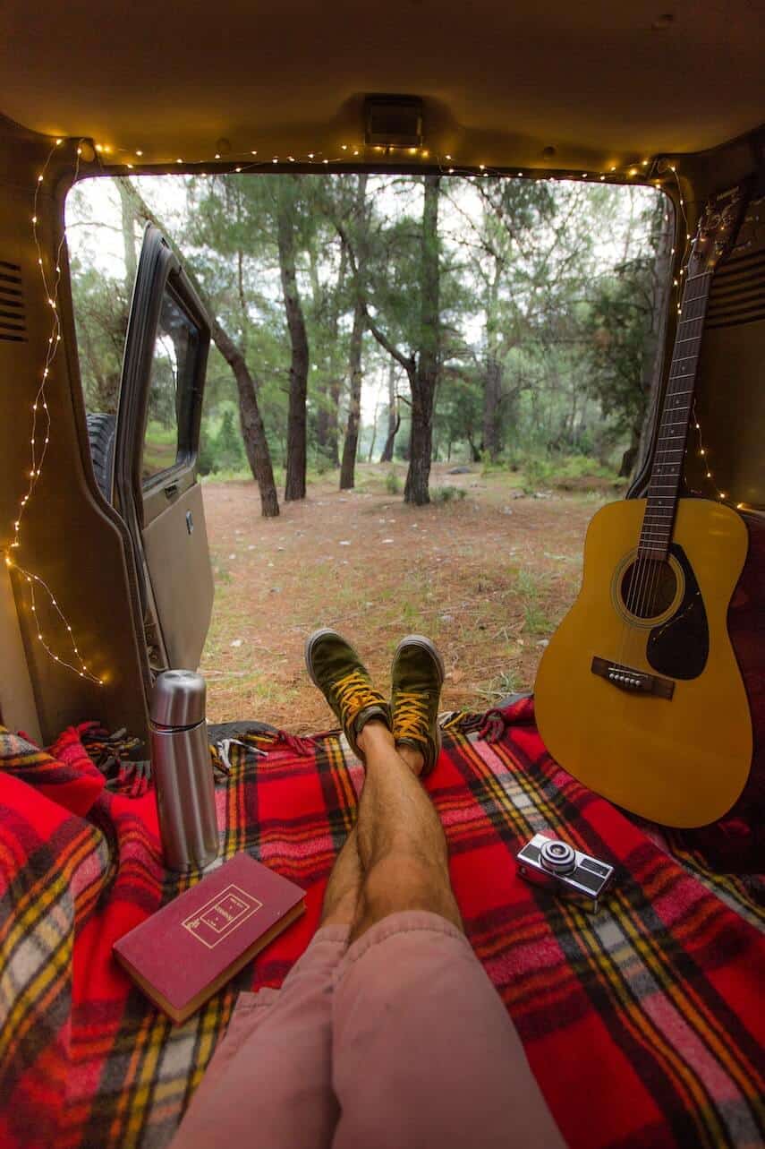 Camervan with the back doors open looking out into the forest. Mans legs wearing pink shorts pointed towards the open doors with a guitar sitting against the side of the vehicle on the right and the door framed by fairy lights