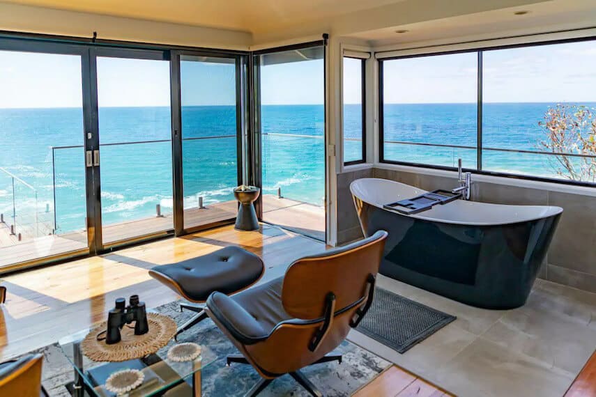 Interior of the Deck House with floor to ceiling windows surrounding the room, a leather chair and coffee table in the centre of the room and free standing black bath on the right