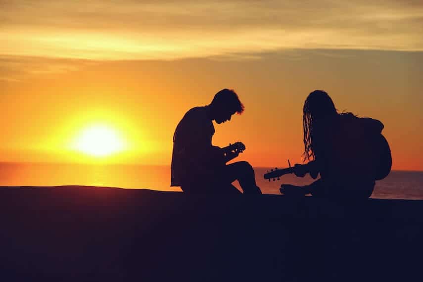 Silhouette on a Couple on the beach with the sun setting behind them. The woman on the right is holding a guitar, the man is sat looking at something in his hands
